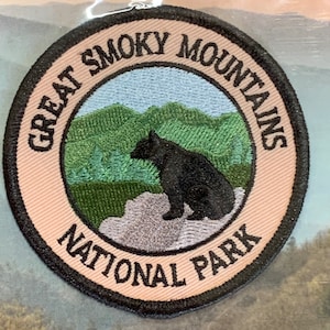 May include: A round, tan embroidered patch with black stitching. The patch features a black bear sitting on a rock in front of a green and blue mountain landscape. The text "Great Smoky Mountains National Park" is written in a black circle around the bear and mountains.
