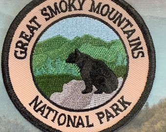 Puede incluir: Un parche redondo, bordado en color beige con costuras negras. El parche presenta un oso negro sentado en una roca frente a un paisaje de montaña verde y azul. El texto "Great Smoky Mountains National Park" está escrito en un círculo negro alrededor del oso y las montañas.