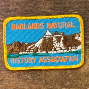 May include: A rectangular patch with a blue background and a yellow border. The patch features an illustration of a mountain range in brown and white, with the text "BADLANDS NATURAL HISTORY ASSOCIATION" in orange.
