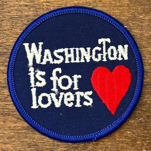 Puede incluir: Un parche redondo azul marino con texto bordado en blanco que dice "Washington is for lovers". Un corazón rojo está en el lado derecho. El parche tiene un borde cosido azul.