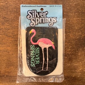May include: Embroidered patch featuring a pink flamingo with a yellow beak, standing in water. The patch is black with the words "Silver Springs" in white and green. The patch is packaged in a clear plastic sleeve, with the text "Embroidered Emblems MADE IN U.S.A."