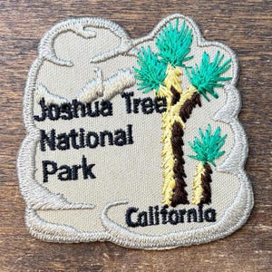 Può includere: Toppa ricamata con la scritta "Joshua Tree National Park California". La toppa ha uno sfondo beige con un albero di Giosuè in marrone, giallo e verde. La toppa ha un bordo decorativo.