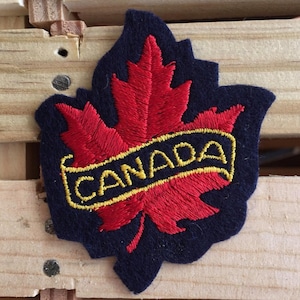 Pode incluir: Um patch de feltro azul marinho em forma de folha de bordo, com um desenho de folha bordada a vermelho. A palavra "CANADA" é bordada a amarelo numa faixa. O patch é provavelmente uma lembrança ou um emblema.