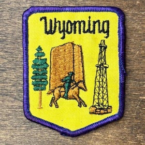 Könnte beinhalten: Ein rechteckiger gestickter Aufnäher mit gelbem Hintergrund und violettem Rand. Das Wort Wyoming ist oben in Schwarz gestickt. Er zeigt eine grüne Kiefer, eine braune Felsformation, eine reitende Person und einen schwarzen Bohrturm.