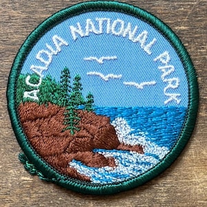 May include: Embroidered patch featuring a coastal scene from Acadia National Park. The design includes a blue sky with white seagulls, a rocky shoreline, and ocean waves. The text "ACADIA NATIONAL PARK" is arched around the top and sides in white.