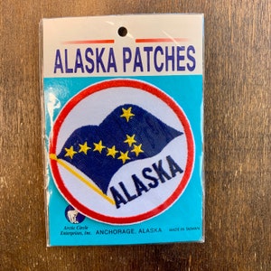 以下が含まれることがあります： 白、赤、青の刺繍が施されたパッチで、「ALASKA」の文字と星がついた青と黄色の旗が描かれています。パッチは、赤い文字と黒い文字で「ALASKA PATCHES」と「ANCHORAGE, ALASKA」と書かれた白いカードに貼られています。カードには、「MADE IN TAIWAN」と「Arctic Circle Enterprises, Inc.」の文字も書かれています。