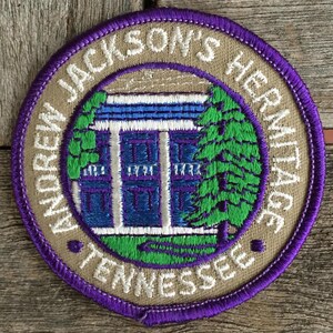 Könnte beinhalten: Ein kreisförmiger gestickter Aufnäher mit beigem Hintergrund und violettem Rand. Die Mitte zeigt ein blau-weißes Gebäude mit weißen Säulen und grünen Bäumen. Der Text lautet Andrew Jackson's Hermitage Tennessee in weißen Buchstaben.