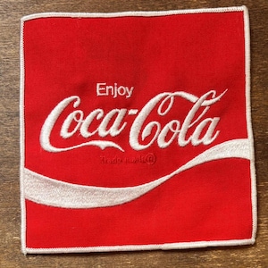 May include: Square red patch with white border featuring the Coca-Cola logo. The words "Enjoy Coca-Cola" are embroidered in white script, with the trademark symbol below. The iconic white wave design is at the bottom.