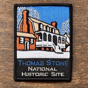 May include: Embroidered patch featuring a detailed illustration of a historic building with a blue sky background. The building is depicted in shades of orange, white, and beige. The text "THOMAS STONE NATIONAL HISTORIC SITE" is embroidered in blue and white.