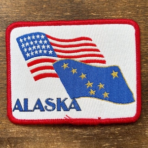 以下が含まれることがあります： アメリカ国旗とアラスカ州旗が刺繍されたパッチ。パッチは白い背景に赤い縁取りがあります。「ALASKA」の文字がパッチの下部に青い文字で刺繍されています。