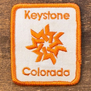Op de afbeelding: Geborduurde patch met de woorden "Keystone Colorado" in oranje letters. De patch heeft een oranje sterrenburst ontwerp in het midden. De patch heeft een oranje rand en is rechthoekig van vorm.