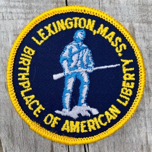 Peut inclure: Un écusson brodé bleu et jaune avec le texte "Lexington Mass. Birthplace of American Liberty" et une silhouette blanche et bleue d'une personne tenant un fusil.
