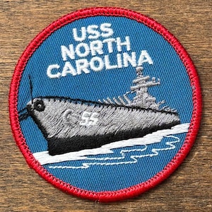 Könnte beinhalten: Runder, gestickter Aufnäher mit dem Schlachtschiff USS North Carolina. Das Design hat einen blauen Hintergrund mit weißem Text und eine detaillierte Illustration des Schiffes. Ein roter Rand umrahmt das Design und schafft eine klassische nautische Ästhetik.