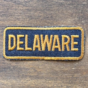 May include: A rectangular patch with the word "DELAWARE" embroidered in gold thread on a navy blue background. The patch has a gold border and is likely designed to be sewn onto clothing or accessories.