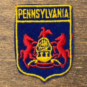 Pode incluir: Um emblema bordado em forma de escudo com fundo azul escuro e borda amarela. A palavra PENNSYLVANIA está bordada em amarelo no topo. O centro apresenta um brasão amarelo com um navio, ladeado por dois cavalos vermelhos e uma águia vermelha.