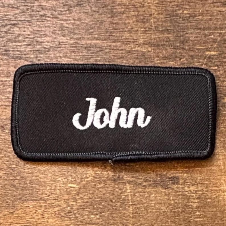 以下が含まれることがあります： 黒い長方形のパッチで、名前「John」が白い筆記体で刺繍されています。パッチには黒い縁取りがあります。背景は木材です。