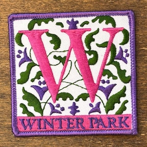 Könnte beinhalten: Gestickter Aufnäher mit weißem Hintergrund, mit einem großen rosa Buchstaben "W" und den Worten "WINTER PARK" in Rosa. Das Design umfasst grüne Blätter und lila florale Akzente, alles eingerahmt von einem lila Rand.