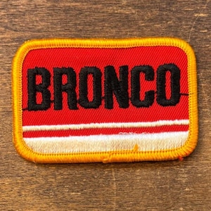 May include: Rectangular patch with the word "BRONCO" in black, embroidered on a red background. The patch has a white and cream stripe at the bottom and a gold border. The patch is likely for a jacket or bag.