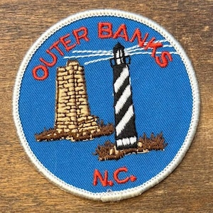 May include: Embroidered patch featuring a blue background with the words "Outer Banks N.C." in red. The patch depicts a lighthouse with black and white stripes and a brown brick structure.