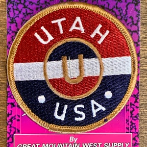 Puede incluir: Un parche redondo bordado con un borde dorado. El parche presenta un diseño rojo, blanco y azul con las palabras "UTAH" y "USA" en letras blancas. El centro del parche presenta una "U" dorada sobre un fondo rojo.