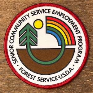 Könnte beinhalten: Runder Aufnäher mit rotem Rand, der den Text "SENIOR COMMUNITY SERVICE EMPLOYMENT PROGRAM" und "FOREST SERVICE U.S.D.A." zeigt. Das zentrale Design umfasst einen Baum, eine Sonne und einen Regenbogen in Grün, Braun, Gelb und Blau.