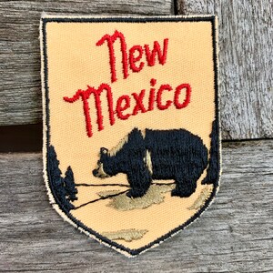 May include: Embroidered patch featuring the text "New Mexico" in red script above a black bear illustration. The bear is set against a tan background with a black border and trees.