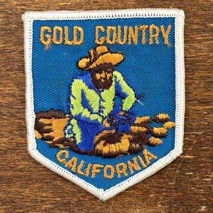 May include: Embroidered patch with a blue background and white border. The patch features a gold miner illustration with the text "GOLD COUNTRY CALIFORNIA" in gold lettering. The miner is wearing a hat and is sitting down.