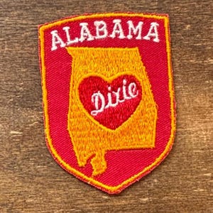 May include: A red, yellow, and white embroidered patch. The patch is shield-shaped with a red border and features the word "ALABAMA" in white block letters at the top. Inside is a yellow outline of the state with a red heart containing the word "Dixie".
