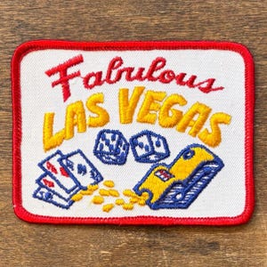 May include: Embroidered patch with a red border and a white background. The patch features the words "Fabulous Las Vegas" in red and yellow, along with images of dice, playing cards, and a slot machine. A fun souvenir.