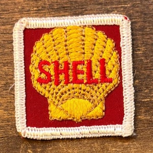May include: Embroidered patch featuring the Shell logo. The design includes a yellow scallop shell with the word "SHELL" in red lettering, set against a red background. The patch is framed by a white stitched border.