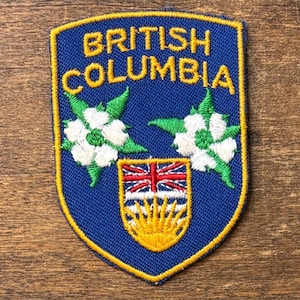 May include: A blue embroidered patch with the words "BRITISH COLUMBIA" in gold. The patch features white and green floral designs, a Union Jack, and a yellow sun emblem. The patch has a yellow border.