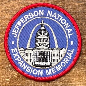 Puede incluir: Un parche circular con un centro azul y un borde rojo. El parche presenta el texto "JEFFERSON NATIONAL EXPANSION MEMORIAL" en blanco, que rodea una ilustración detallada de un edificio con una cúpula.