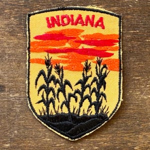 May include: Embroidered patch with the word "INDIANA" in red, above an orange and red sunset. Black corn stalks are embroidered below against a yellow backdrop, with a black border.