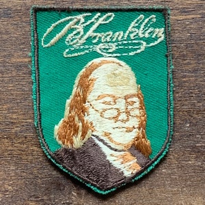 May include: A green embroidered patch with a portrait of Benjamin Franklin and his signature "B Franklin" in white thread.
