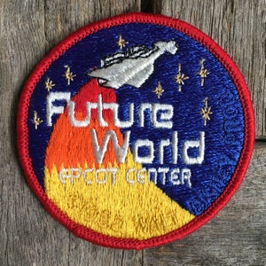 May include: A blue and yellow embroidered patch with a spaceship flying over a mountain. The text "Future World EPCOT Center" is embroidered on the patch.