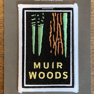 May include: A white embroidered patch with a black background. The patch features a stylized image of trees with green and orange details. The text "MUIR WOODS" is written in yellow letters.