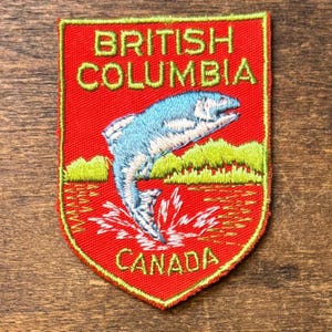 May include: Embroidered patch with a red background, featuring a leaping blue fish above green hills and water. The text "BRITISH COLUMBIA CANADA" is in yellow. The patch has a yellow border.