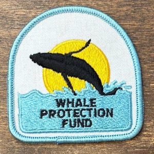 May include: A semi-circular embroidered patch featuring a black whale silhouette leaping in front of a bright yellow sun. Below the whale, light blue waves are depicted with the black embroidered text WHALE PROTECTION FUND centred.