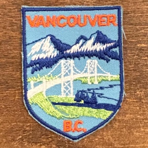 May include: Embroidered patch with a shield shape, displaying the word "VANCOUVER" in orange. The design includes a bridge, mountains, and the text "B.C." at the bottom, set against a light blue backdrop.