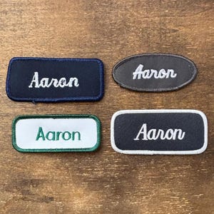 May include: Four embroidered name patches, each with the name "Aaron" in white script. The patches are in different shapes: a navy blue rectangle, a grey oval, a white rectangle with a green border, and a black rectangle with a white border.