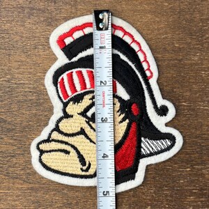Pu&ograve; includere: Una toppa ricamata raffigurante una mascotte che indossa un elmo con dettagli rossi e neri, posizionata accanto a un metro a nastro bianco che mostra un'altezza di circa 11,5 cm. Il disegno utilizza fili di colore beige, rosso, nero e bianco.