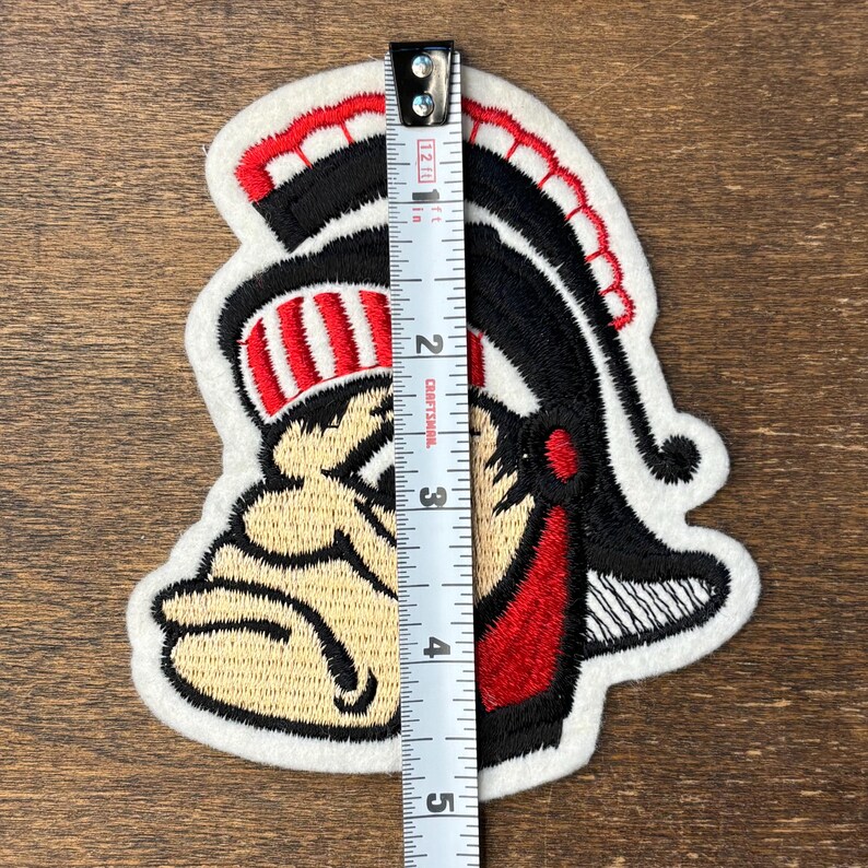 Pu&ograve; includere: Una toppa ricamata raffigurante una mascotte che indossa un elmo con dettagli rossi e neri, posizionata accanto a un metro a nastro bianco che mostra un'altezza di circa 11,5 cm. Il disegno utilizza fili di colore beige, rosso, nero e bianco.