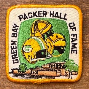 May include: A square embroidered patch with a yellow border featuring the text GREEN BAY PACKER HALL OF FAME in black. The center displays two yellow football helmets over a green background, with a tan building illustration at the bottom.