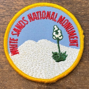 May include: Embroidered patch with the text "WHITE SANDS NATIONAL MONUMENT" in red on a light blue background. The patch features a white mountain range and a green plant with white flowers. The patch has a yellow border.