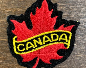 May include: An embroidered patch featuring a vibrant red maple leaf centred on a black background. A yellow banner with a black border is stitched across the middle of the leaf, displaying the word CANADA in bold, yellow, capitalised letters.