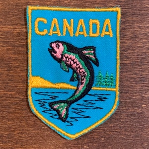May include: A blue and yellow embroidered patch with the word "CANADA" in yellow lettering and a pink and green fish jumping out of blue water.