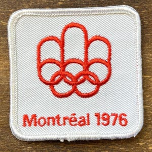 Puede incluir: Un parche cuadrado bordado en blanco con los anillos olímpicos rojos y el texto "Montréal 1976". El parche tiene un borde blanco. Conmemora los Juegos Olímpicos de Verano de 1976.