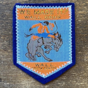 Könnte beinhalten: Ein blau-orange-weiß gestickter Aufnäher mit einem Cowboy auf einem buckenden Bronco. Der Aufnäher trägt oben die Aufschrift "WE MADE IT" und "WALL DRUG" und unten "WALL SOUTH DAKOTA". Der Aufnäher hat einen blauen Rand.