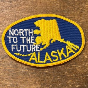Könnte beinhalten: Gestickter Aufnäher mit blauem Hintergrund und gelbem Rand. Der Aufnäher zeigt eine gelbe Umrisslinie von Alaska und den Text "NORTH TO THE FUTURE ALASKA" in Weiß und Gelb.