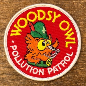 Op de afbeelding: Geborduurde patch met een rode achtergrond en witte rand. De patch toont een cartoon uil karakter in oranje, groen en geel, met de woorden "WOODSY OWL POLLUTION PATROL" in geel en wit.